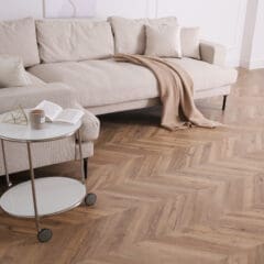 A cozy living room with a beige sectional sofa, two pillows, and a tan throw blanket. A round side table with a candle, open book, and geometric decor sits on a herringbone wood floor. Designed by our home renovations team, soft neutral tones create a serene atmosphere.