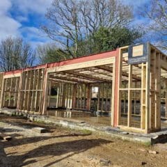 A wooden frame of a house under construction on a concrete foundation, surrounded by trees, highlights the expertise of our building company. The sky is partly cloudy, and construction materials are scattered around the site, showcasing a step towards your dream home.