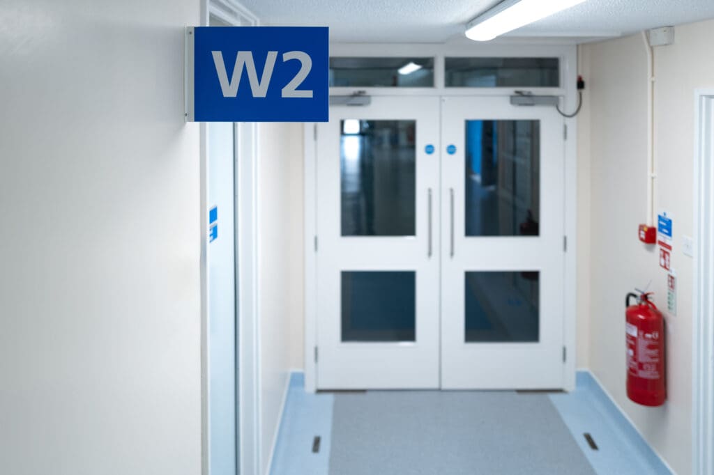 A hallway with white walls and double doors at the end. A blue sign with "W2" hangs from the ceiling. A red fire extinguisher is mounted on the right wall near the doors, adding a touch of practicality often seen in commercial renovations.